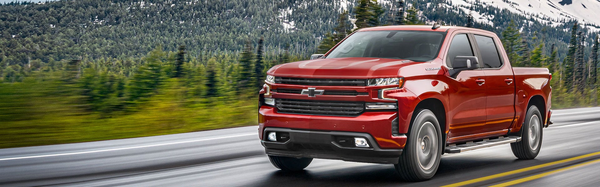 A bold red Chevrolet Silverado pickup truck is driving on a scenic highway, surrounded by lush green forests and snow-capped mountains in the background.
