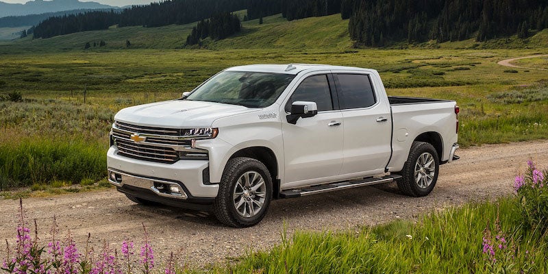 A silver Chevrolet Silverado drives through a picturesque countryside road, surrounded by green fields and wildflowers, showcasing its versatility.