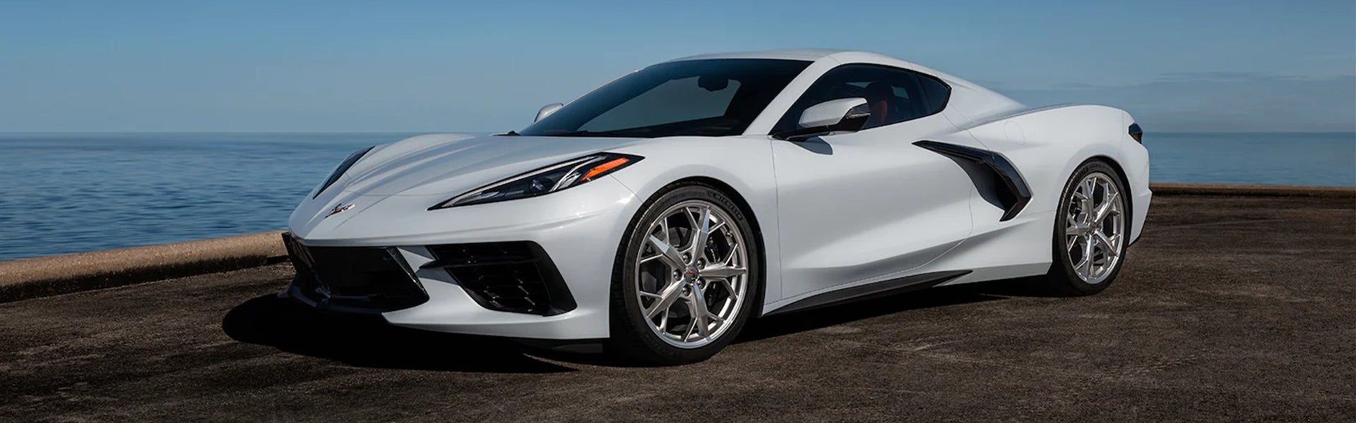 White Chevrolet Corvette parked near a coastal backdrop with a clear blue sky.