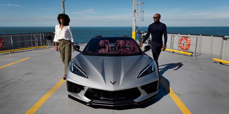 A silver Chevrolet Corvette convertible with two elegantly dressed individuals on a ship deck.