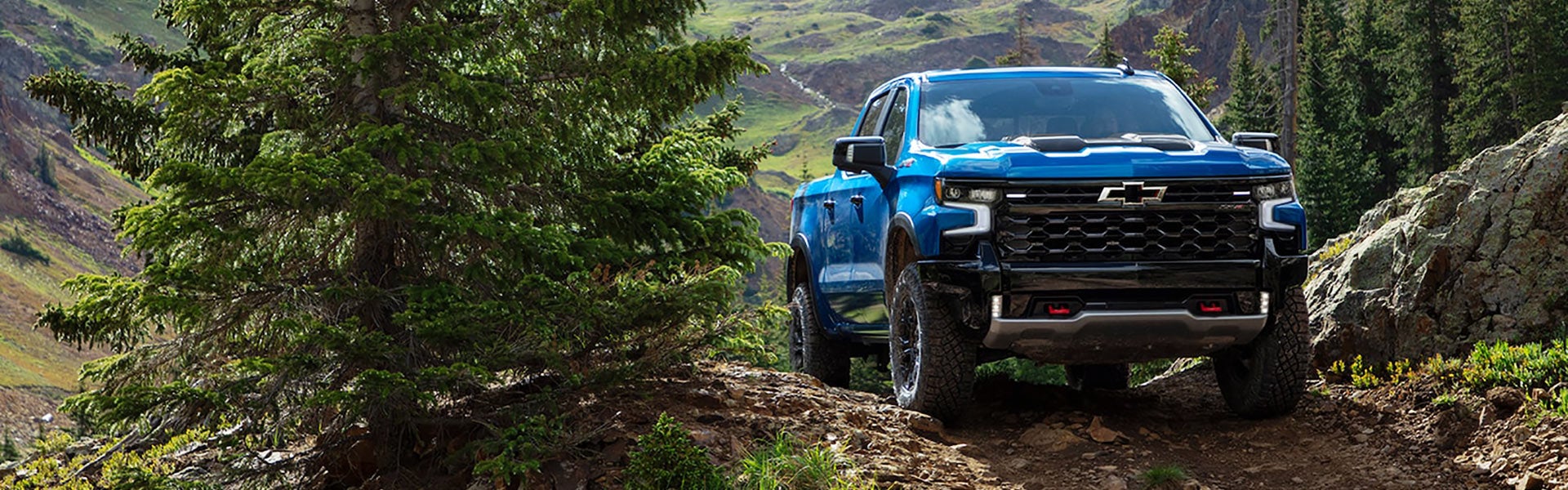 A blue Chevrolet Silverado ZR2 pickup truck navigating a rugged off-road trail in a mountainous landscape, surrounded by greenery and rocky terrain.